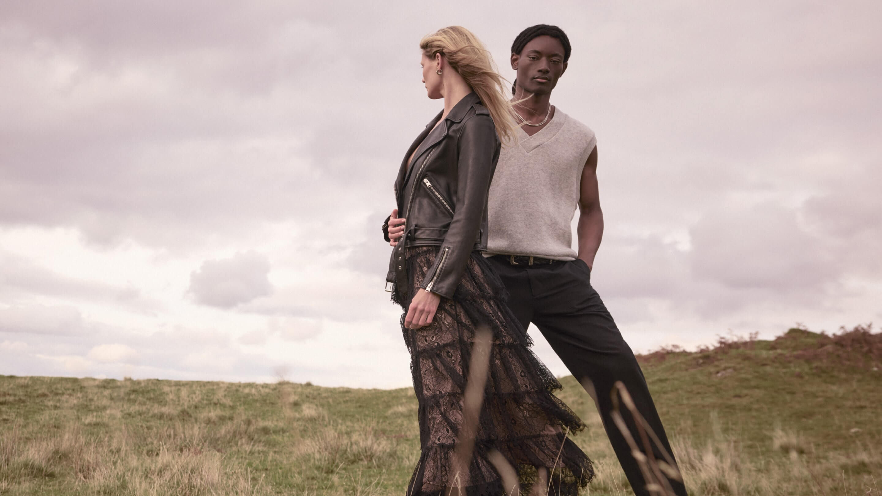 A man and woman in a grass field wearing white knitwear with black jeans and leather jacket over a maxi dress